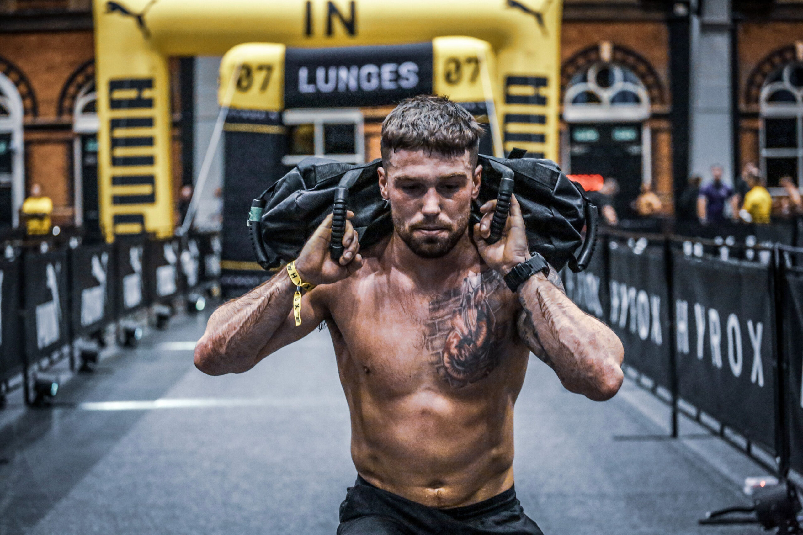 Mann absolviert Sandbag Lunges im HYROX-Wettkampf – Fokus, Balance und Durchhaltevermögen.