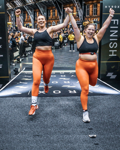 Zwei HYROX-Athletinnen im Double Women Format überqueren gemeinsam jubelnd die Ziellinie.